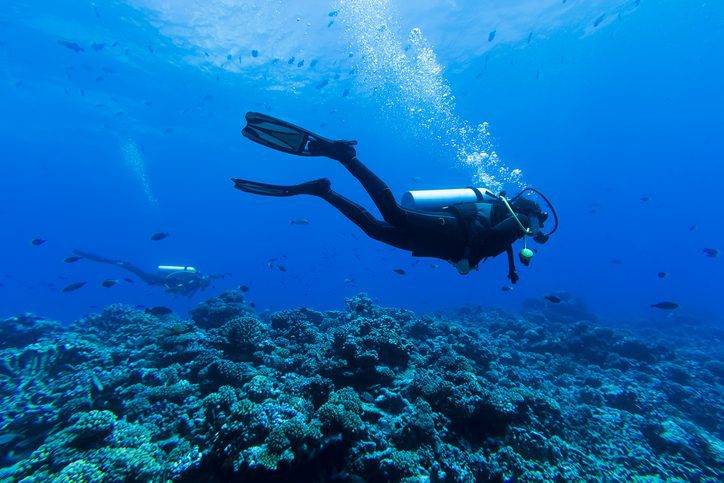 A DSLR underwater photo of a diver at Tiputa Pass, Rangiroa, French Polynesia. She is wearing black swimsuit and is exhaling bubbles. The diver is swimming over the reefs surrounded by the blueish of the sea . At the backgroung there is another diver swimming in the same direction. There are some small fishes all around.